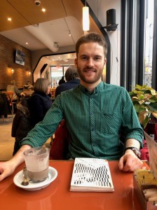 Author photo: Facing camera sitting in a restaurant with hot chocolate and a book of poems in front of him, smiling. 