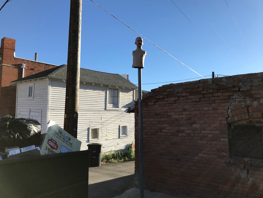Photo of a store mannequin, female, outside in the back alley, near garbage. Old brick and old wood houses, Butte, Montana. 