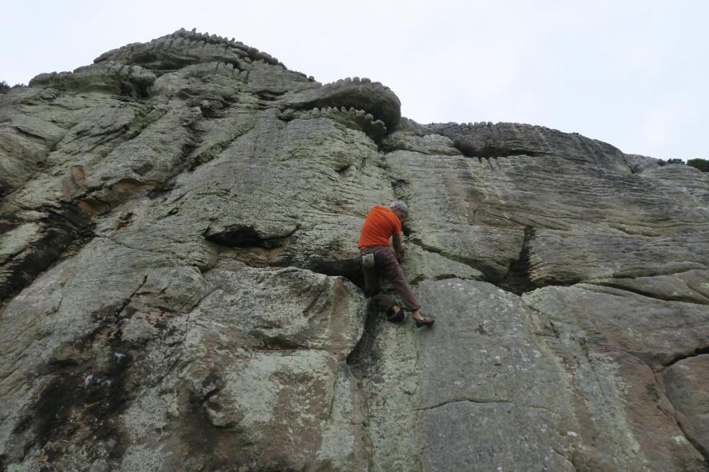 Photo of the author by Nikki Clayton (2023). Rock climbing almost sheer rocks, photo angle upward to the ridge and the sky.
