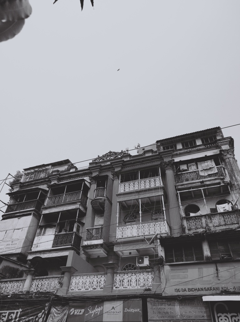 Black and white photo of an old apartment building, colonial style, address 136/3A Bidhan Sarani. At bottom are awnings for shops. Ornate pillars and balconies with metal patterned fronts.