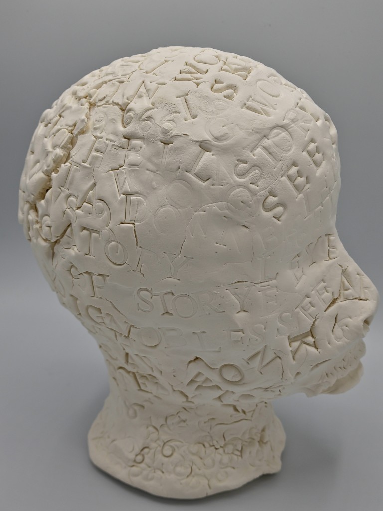 Clay Head #2 in right profile. 
text includes gouged stamped words: STORY   SEE KNOW repeated and scattered over the clay head.