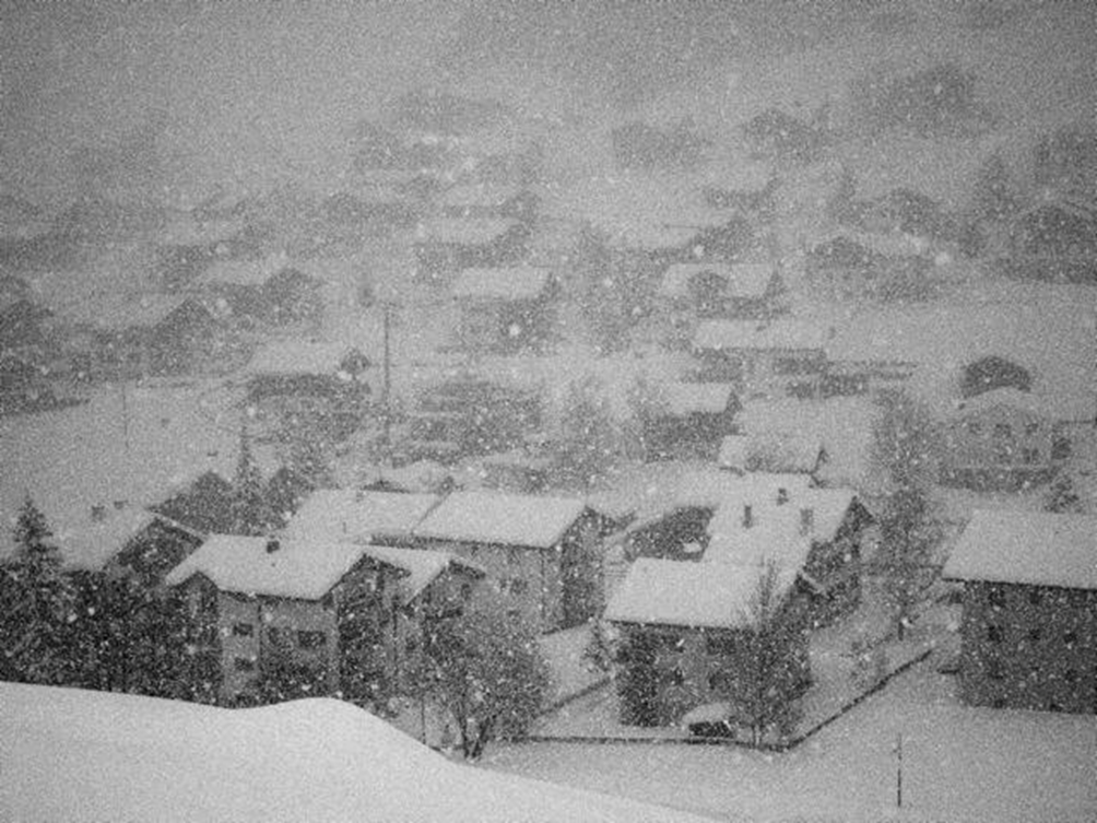 A photo looking down on a small snowbound complex of low rise apartment houses dotted around a hill; there is a uniform quality also something dreamy and otherworldly, thick snow;   falling, black and white - 