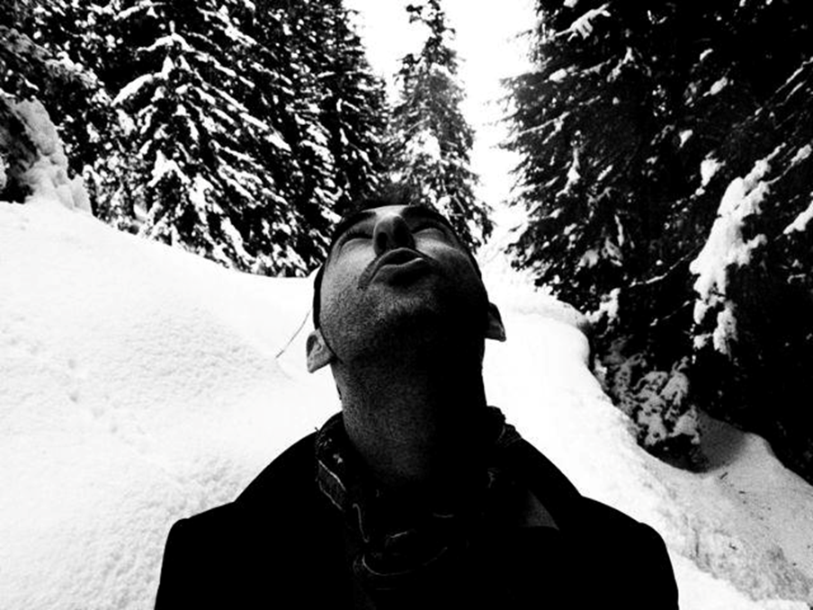A portrait of a young man looking up into a snowy wintry sky, evergreen trees behind him . Black and white.